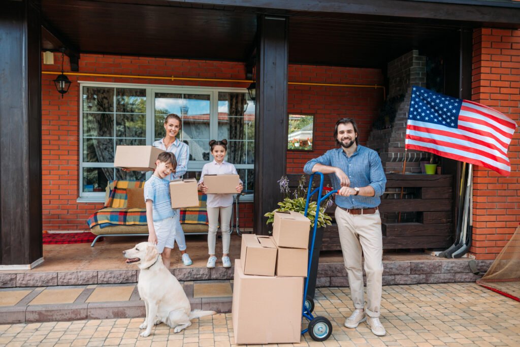 Famille avec enfants et chien labrador devant des cartons de déménagement empilés à l'entrée d'une maison avec drapeau américain